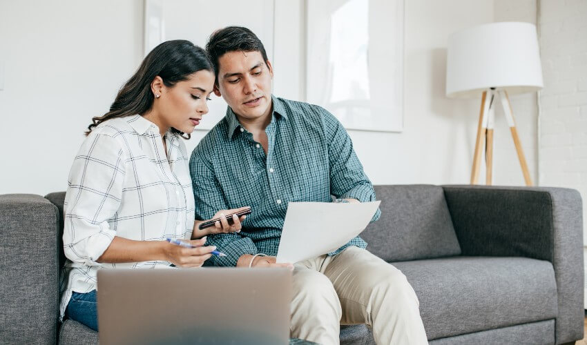 Young couple sitting on couch looking at finances with calculator, laptop and spreadsheet.